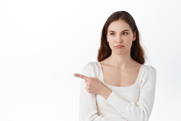 Недовольная и скептически настроенная молодая женщина, указывающая пальцем в сторону и смотрящая влево с недовольным и невпечатленным лицом, стоящим на белом фоне