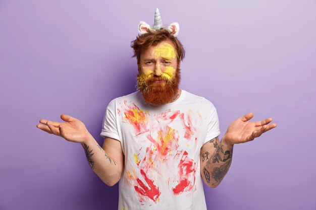 Ничего не подозревающий бородатый молодой человек, испачканный разноцветными акварельными красками, нерешительный разводит руки, носит рог и уши единорога, белую футболку с пятнами, изолирован на фиолетовой стене
