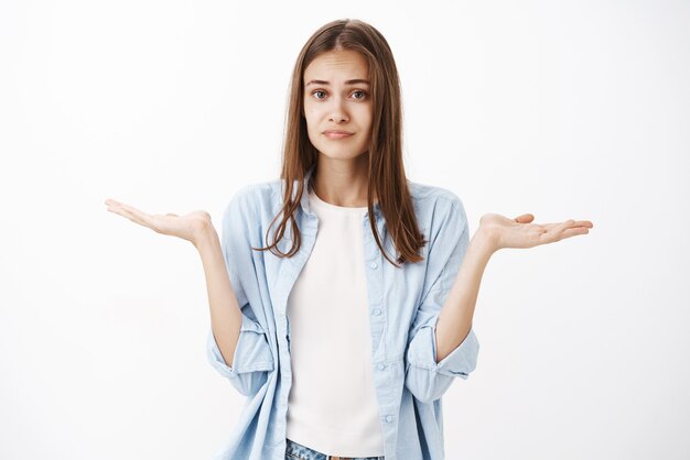 気づかず、無知で魅力的なフェミニンな女の子。青いブラウスに茶色の髪をし、肩の近くに上げられた手で肩をすくめて、はっきりしない残念な表情の白いtシャツ