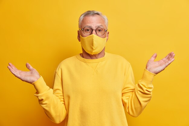 unaware grey haired man doesnt know how what happening spreads palms and stands confused against yellow vivid wall wears protective mask during coronavirus pandemic