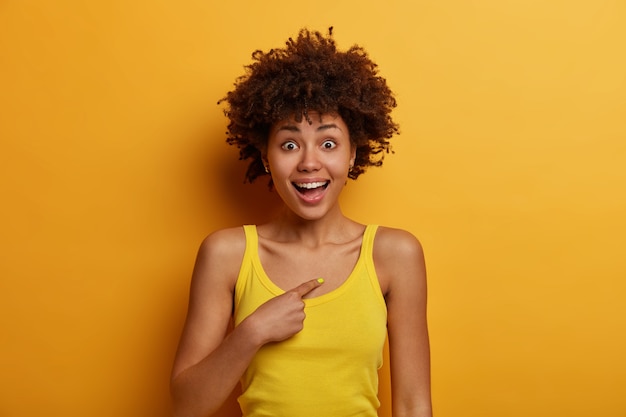 Unbelievable, really me? Surprised glad dark skinned woman points at herself, shocked and happy to hear her name or being chosen, becomes team member, dressed in casual clothes, poses over yellow wall