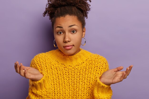 Uncertain adult woman with curly combed hair, shruggs shoulders, keeps palms sideways, has questioned perplexed face expression