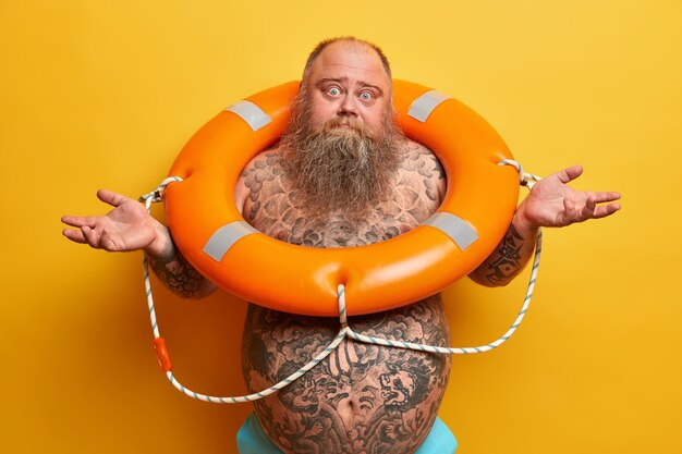 Неуверенный бородатый мужчина с большим татуированным животом, разводит руки в стороны, чувствует себя неуверенно и нерешительно, стоит в оранжевом спасательном круге, учится плавать, изолирован на желтой стене. Время для купания