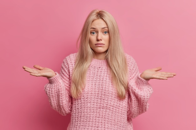 不確かな金髪の女性が手のひらを広げ、屋内で疑わしい立っている2つのオプションのどちらかを選択することはできませんニットの暖かいセーターを着ています