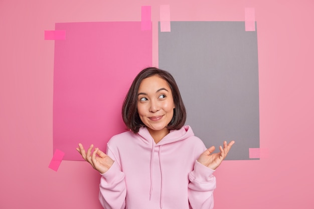 Uncertain hesitant Asian woman spreads palms feels unaware thinks about choice has no idea being clueless stands questioned wears casual hoodie poses against blank empty wall