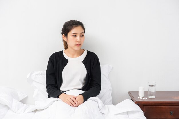 An uncomfortable woman sits on the bed and has medicine on the table.