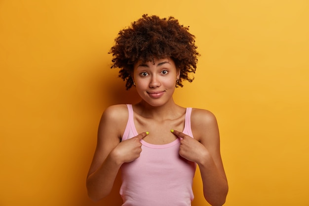 Unconfident hesitant curly haired adult woman points at herself, asks doubtfully whether she is good candidate, dressed casually, isolated over yellow wall, looks friendly, asks question