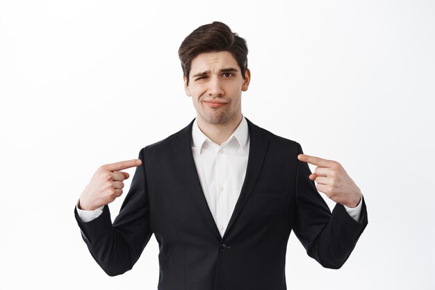 Unconfident and skeptical man in black formal suit points at himself frowns and look doubtful stands against white background