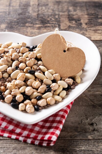 Uncooked assorted legumes in heart shape plate on rustic wooden table.