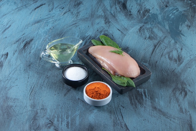 Uncooked chicken breast and spinach on a wooden plate next to spice, salt and oil bowls , on the blue surface.