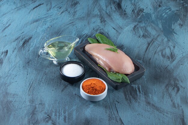 Uncooked chicken breast and spinach on a wooden plate next to spice, salt and oil bowls , on the blue surface.