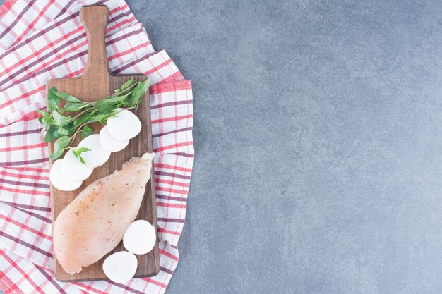 Uncooked chicken fillet with radish slices on wooden board.