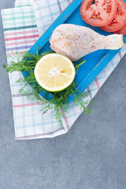 Uncooked chicken leg, tomato and lemon slice on blue plate.