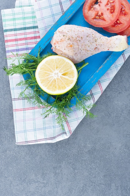 Uncooked chicken leg, tomato and lemon slice on blue plate.