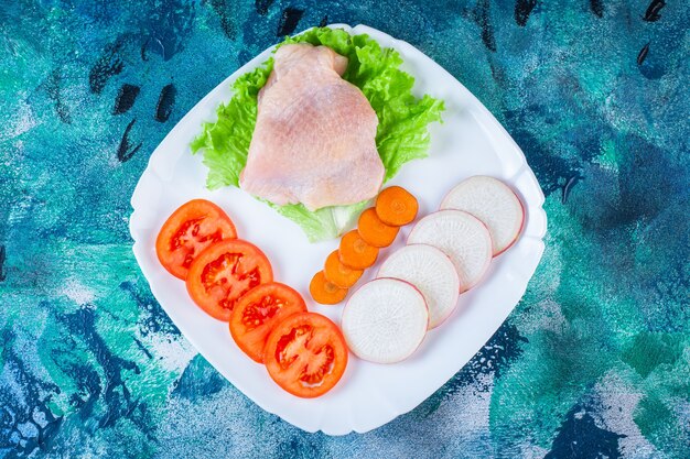 Uncooked chicken meat next to tomatoes ,radish and carrot on a plate