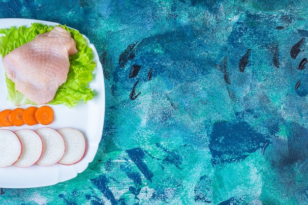 Uncooked chicken meat next to tomatoes ,radish and carrot on a plate