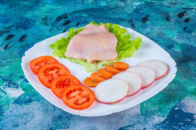 Uncooked chicken meat next to tomatoes ,radish and carrot on a plate