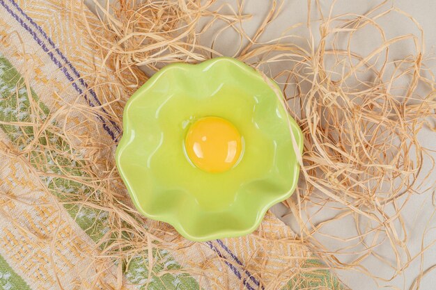 Uncooked egg in green plate with hay on marble table.