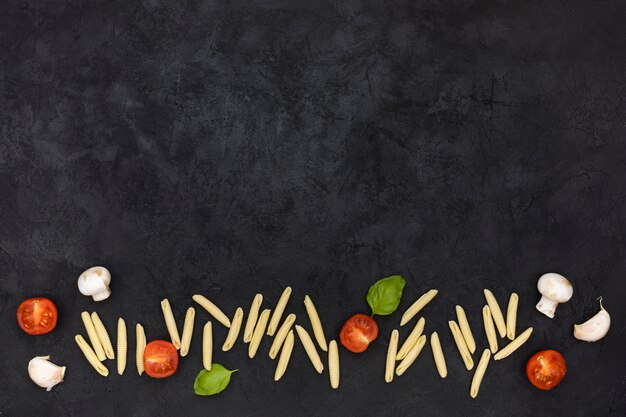 Сырая паста гарганелли с половинками помидоров; гриб; зубчик чеснока и базилик на дне черного текстурированного фона