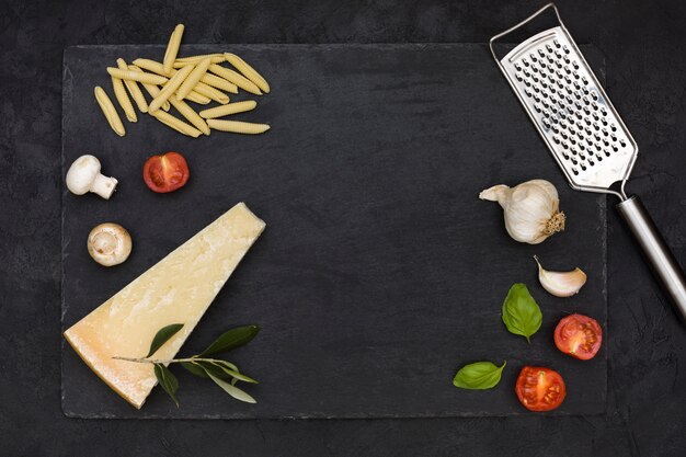 Uncooked italian garganelli pasta with ingredients and cheese grater on rock slate against black backdrop