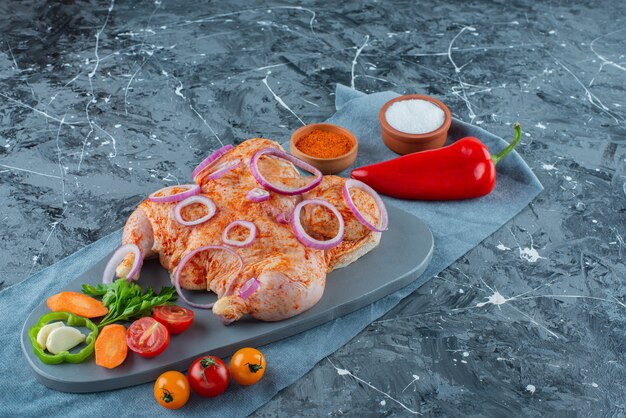 Pollo marinato crudo con verdure su una tavola su un pezzo di tessuto, sullo sfondo blu.