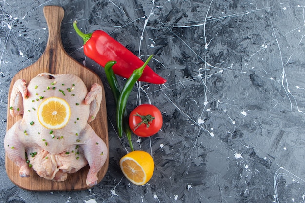 大理石の背景に、野菜の横にあるまな板の上で未調理のマリネした丸ごとの鶏肉。