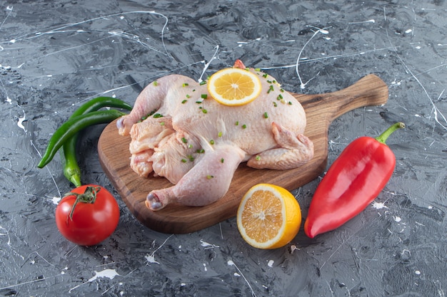 Uncooked marinated whole chicken on a cutting board next to vegetables , on the marble surface.