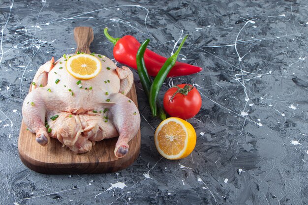 Uncooked marinated whole chicken on a cutting board next to vegetables , on the marble surface.