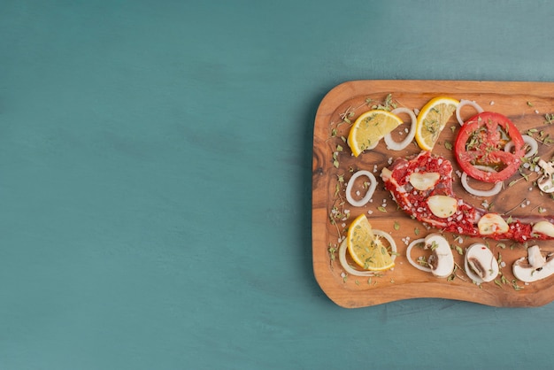 Uncooked meat piece with vegetables on blue table.