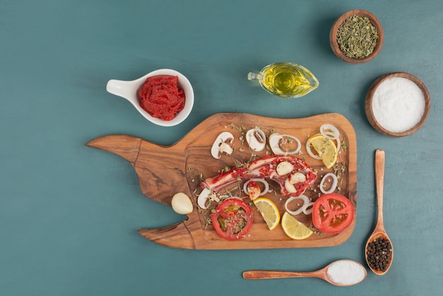 Uncooked meat piece with vegetables, oil and spices on blue table.