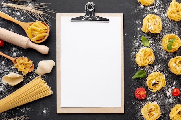 Uncooked pasta assortment with flour on black background with clipboard mock-up