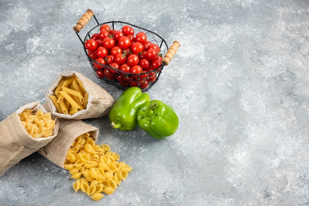 Uncooked pastas in a rustic bag with cherry tomatoes and peppers on marble table.