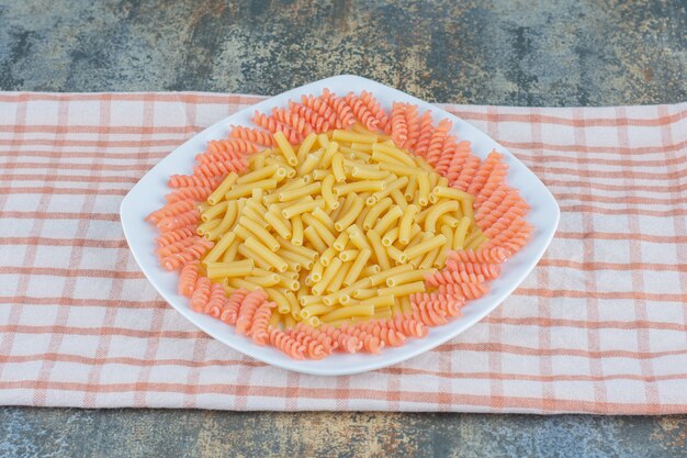 Uncooked penne and fusilli pasta in bowl on towel, on the marble surface. 