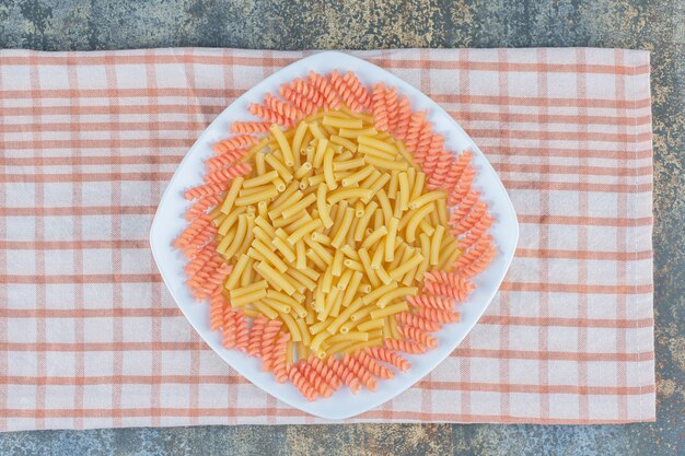 Uncooked penne and fusilli pasta in bowl on towel, on the marble surface. 