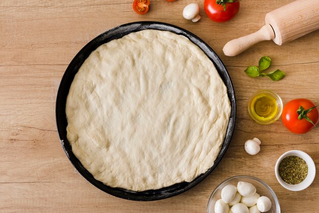 Сырое тесто для пиццы с начинкой и скалкой на деревянном фоне