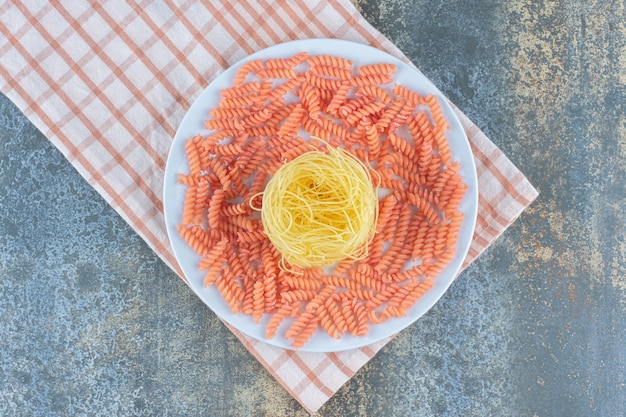 Сырые спагетти и макароны фузилли в миске на полотенце на мраморном фоне.