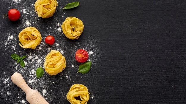 Uncooked tagliatelle on black background with tomatoes rolling pin and copy space