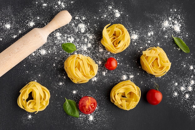 Uncooked tagliatelle on black background with tomatoes and rolling pin