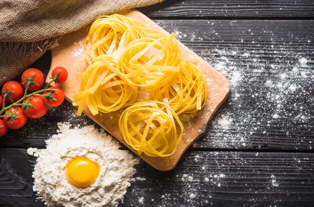 Uncooked tagliatelle on chopping board with egg york in flour and tomatoes on wooden plank