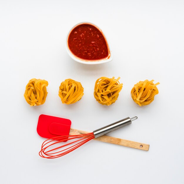 Uncooked tagliatelle pasta and tomato sauce with kitchen utensil isolated on white background