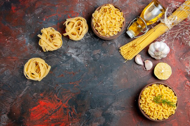 Uncooked three spaghetti and butterfly pastas in a brown bowl and green onion lemon garlic oil bottle on mixed color background