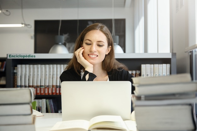 Undergraduate student woman preparing for exams, working on laptop, using wireless Internet connection while sitting at desk with huge stacks of books at college library, resting her elbow on table