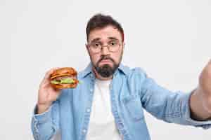 Бесплатное фото Несчастный бородатый взрослый европейский мужчина ест мусор, держит вкусный гамбургер, делает селфи кошельки, губы недовольны, выражение лица носит круглые очки, джинсовую рубашку