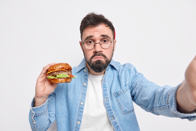 불행한 수염 난 성인 유럽인 남자가 정크를 먹고 맛있는 햄버거를 먹고 셀카 지갑을 만듭니다 입술에 불만이 있습니다 표정이 둥근 안경 데님 셔츠를 입습니다.