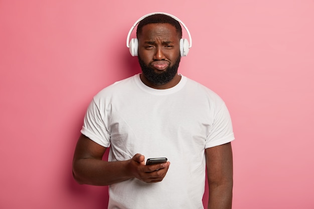 Unhappy black unshaven man listens music in headphones, has displeased expression, holds mobile phone, dressed casually, upset as cannot download song in playlist