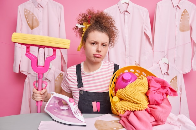 Unhappy bored housemaid doesnt want to do housework holds mop for cleaning floor in room stands near ironing board with basket of laundry dressed casually has gloomy expression. Domestic work