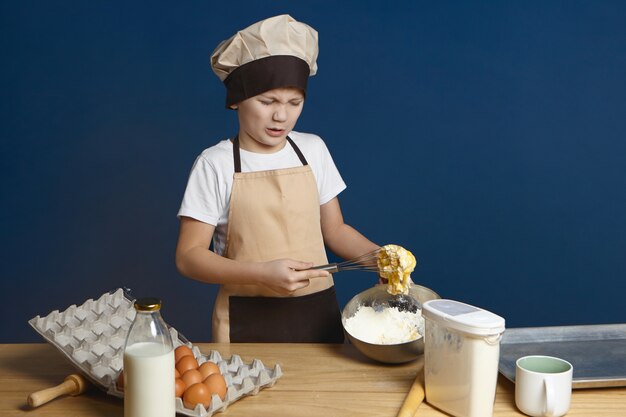 Unhappy Caucasian little boy in chef uniform standing at kitchen table and frowning having disgusted upset expression