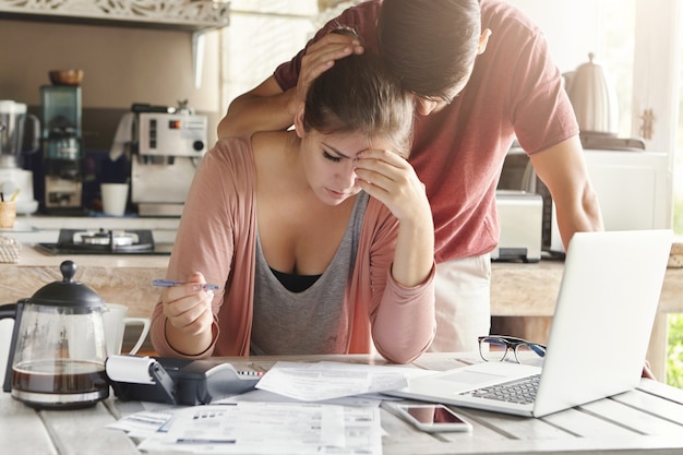 Unhappy couple unable to pay loan on time: stressed female doing paperwork sitting at table with laptop, papers, calculator and cell phone. Man trying to support his wife