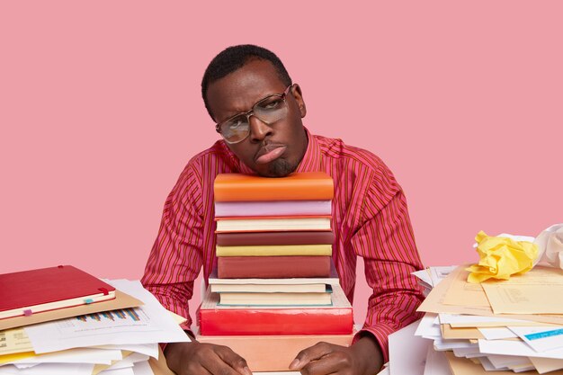 Unhappy dark skinned man in formal clothes, being disappointed, tired of work, leans on pile of books, wears big spectacles and formal shirt