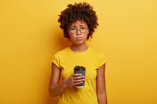 Unhappy dark skinned student holds disposable cup of caffeine beverage